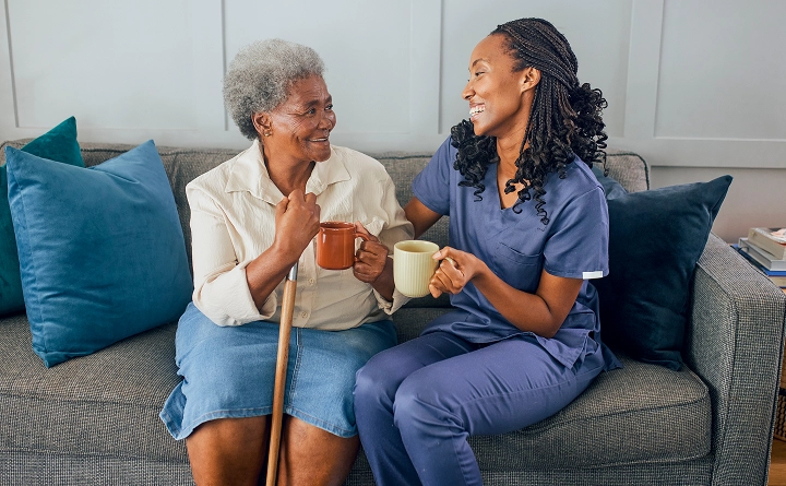 senior woman with cane and nursing professional sitting on a couch with mugs